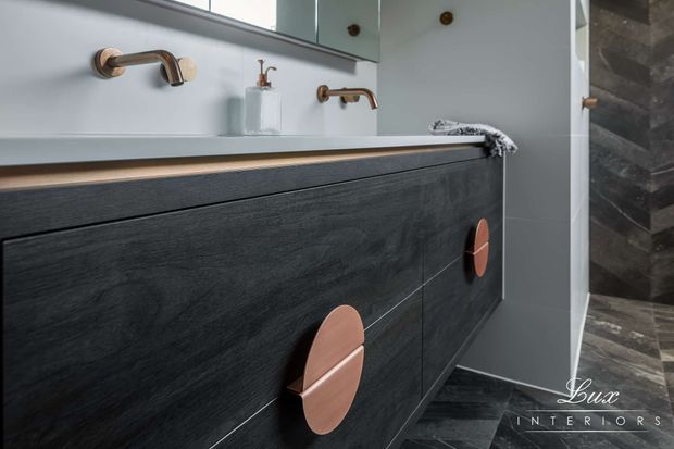 Close-up view of a refined contemporary bathroom vanity with dark timber-look cabinetry, slim pale benchtop, round copper-toned handles, wall-mounted copper-toned tapware, and a large mirror. Dark patterned wall and floor tiles add contrast, while soft grey walls create a restrained, luxurious feel.