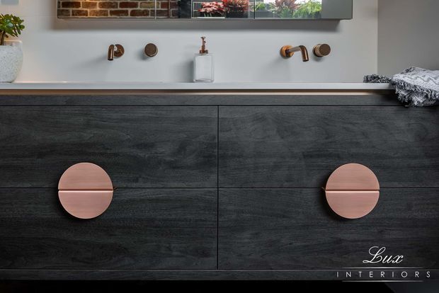 Close-up of a contemporary bathroom vanity with a long pale benchtop, dark charcoal wood-grain cabinetry, and two round blush-pink drawer pulls. Two wall-mounted copper-toned tap sets and a white soap dispenser sit against a light wall, with a mirrored cabinet above.