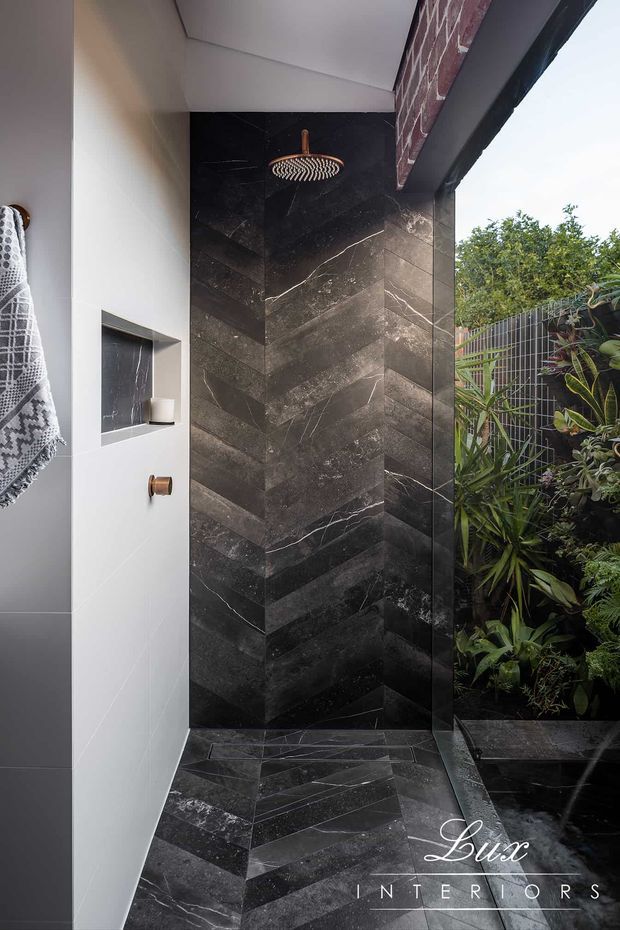 A sophisticated open-air shower enclosure featuring dramatic dark veined stone-look wall and floor tiling arranged in a chevron pattern. A bronze rainfall showerhead and controls contrast with the black surfaces, while a recessed wall niche provides storage. The shower opens directly onto a lush planted courtyard, with a textured brick exterior detail overhead and a dark metal privacy screen.