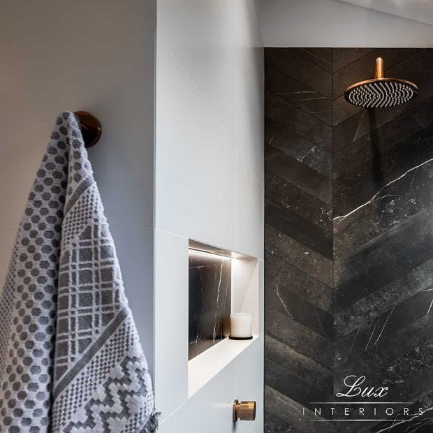A contemporary bathroom shower area featuring a dark veined stone-look tiled feature wall in a chevron arrangement, a copper-toned rainfall showerhead and controls, a recessed illuminated niche, and light neutral walls. A patterned towel hangs from a round wall hook in the foreground.