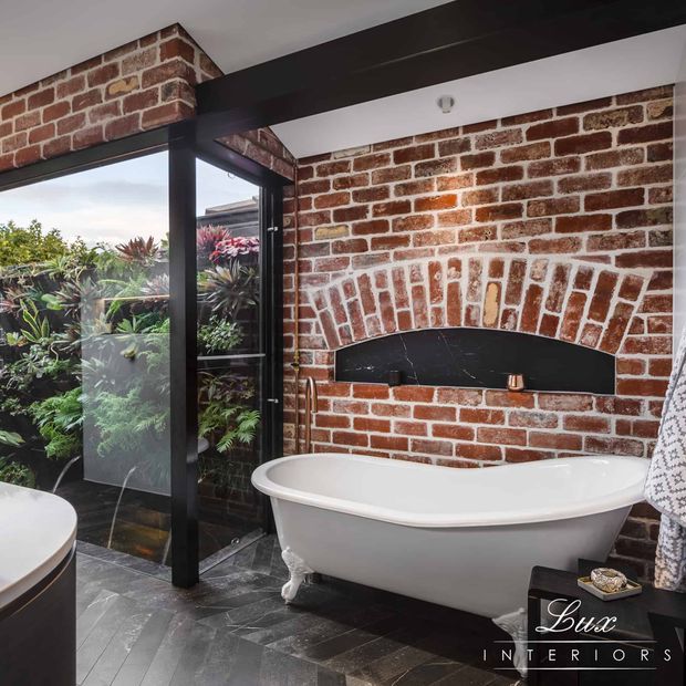 A luxurious bathroom featuring a freestanding white bathtub set against an exposed red-brick wall with a striking arched black fireplace-like recess. Black-framed glazing opens to a lush planted courtyard, while dark slate-style floor tiles, black structural beams, and a patterned towel add contrast.