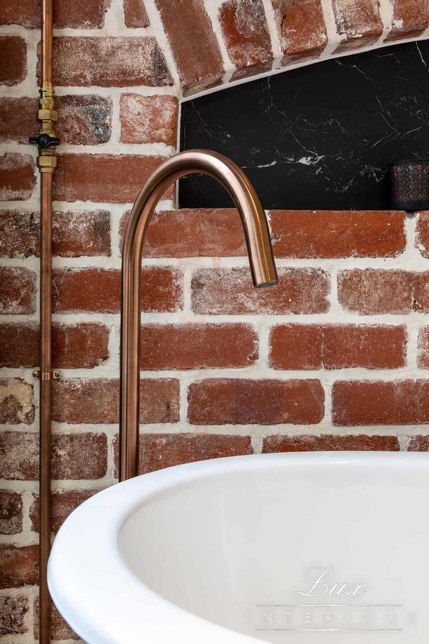 Close-up of a freestanding white bathtub with a tall curved copper-toned wall-mounted faucet and exposed copper pipework. The backdrop is a rustic red brick wall with white mortar, a black marble-look splashback panel, and a small dark patterned wall detail. The image has a warm, industrial bathroom aesthetic.