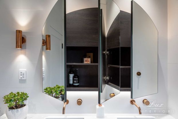 A refined bathroom vanity area with a white built-in mirrored cabinet featuring curved doors, dark timber shelving, and integrated storage. Warm brass or copper wall sconces and tapware contrast with the white tiled or painted surfaces, while a small potted plant adds greenery. The mirrored doors reflect the vanity and cabinetry.