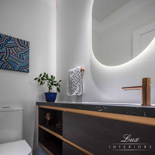 A contemporary powder room with a wall-mounted dark timber vanity, black marble-look benchtop, brushed copper tapware, white tiled walls, a large rounded backlit mirror, and a white toilet. A patterned artwork and folded towel add decorative detail.