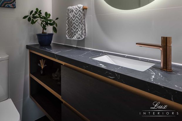 Contemporary bathroom vanity with a dark marble-look benchtop, integrated white basin, black timber-look cabinetry with warm wood edging, and a brushed bronze wall-mounted tap. Large-format grey wall tiles, a round mirror, patterned towel, and potted plant complete the refined scheme.