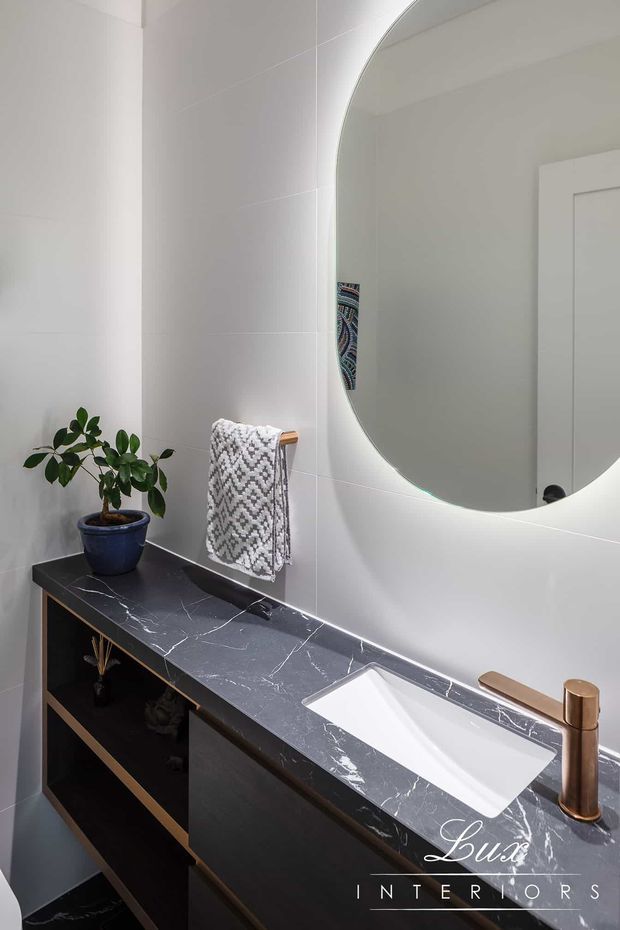 A bright, contemporary bathroom vanity with a large rounded-rectangle mirror, white tiled walls, dark marble-look benchtop, integrated white basin, dark cabinetry, and a warm bronze tap. A small potted plant and patterned hand towel add subtle decorative detail.