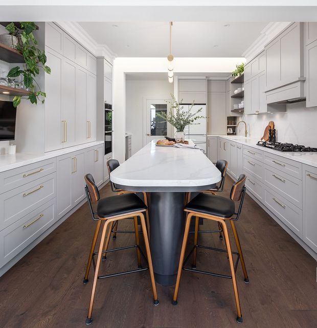 Bright, spacious contemporary kitchen with extensive pale grey built-in cabinetry on both sides, brass-toned hardware, dark timber flooring, and a large white stone island with a sculptural dark base. Several black and timber bar stools surround the island, while pendant lights, open shelving, integrated appliances, and indoor greenery complete the refined interior.