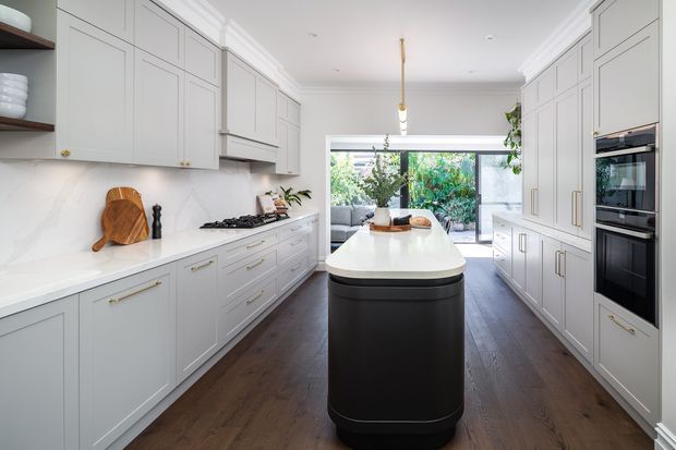 Bright, spacious contemporary kitchen with symmetrical full-height pale cabinetry, a large dark island with a white stone benchtop, integrated double ovens, a marble-look splashback, dark timber flooring, and a linear pendant light. Glazing at the rear opens the kitchen visually to a leafy outdoor area and adjacent living space.