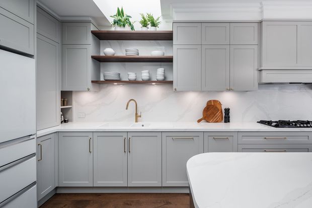 Bright, refined kitchen with grey Shaker-style cabinetry, white marble-look splashback and benchtops, open dark timber shelving displaying white crockery, brass tapware, integrated refrigerator, gas cooktop and a curved island in the foreground. Potted greenery adds a natural accent.