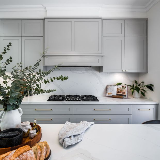 A refined grey-and-white kitchen with classic Shaker-style cabinetry, a marble-look backsplash and benchtops, brass hardware, and a built-in gas cooktop beneath a concealed rangehood. Decorative greenery, a cookbook, bread, and serving items add warmth to the otherwise light, polished space.
