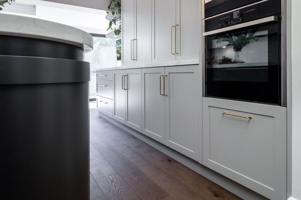 A bright, refined kitchen interior featuring extensive white shaker-style cabinetry with long brass handles, integrated black appliances, dark timber flooring, and a prominent rounded black-and-white island or peninsula in the foreground. Greenery is visible near the upper cabinetry, adding a natural accent.