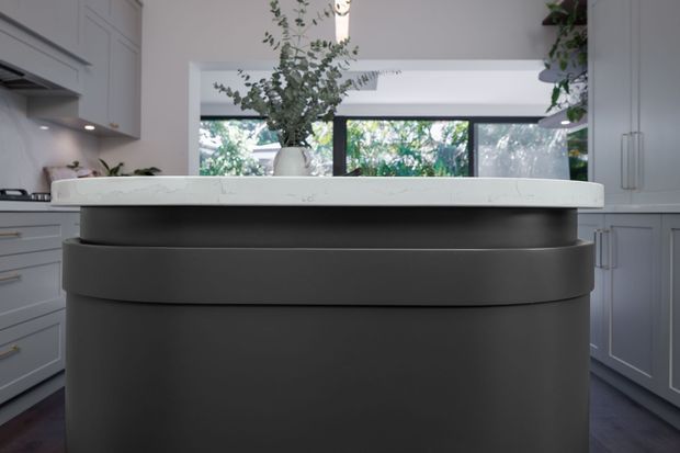 A refined kitchen centred on a large curved dark island with a thick white stone benchtop. Pale grey cabinetry with recessed panel doors and slim metal handles surrounds the space, while black-framed windows provide views to leafy greenery. A vase of foliage sits on the island, and subtle under-cabinet lighting adds warmth.