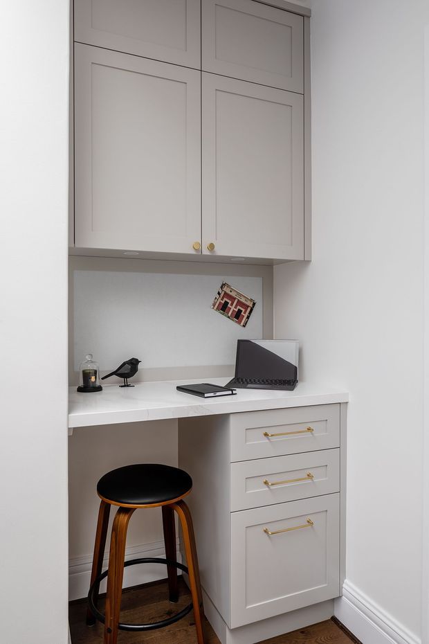 Compact built-in study nook with a white paneled overhead cabinet, white desk surface, and matching drawer cabinetry with brass pulls. A laptop and small decorative objects sit on the desk, with a black-and-wood stool tucked underneath. The nook is set into a light neutral wall with timber flooring and white skirting boards.