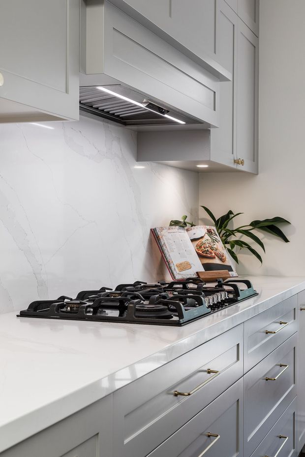 Close-up photograph of a refined, light-toned kitchen cooking zone. A large black gas cooktop sits within a white veined stone benchtop and matching full-height splashback, beneath a stainless-steel rangehood concealed by pale grey shaker-style cabinetry. Brass pulls and knobs provide subtle contrast, with a cookbook and plant on the rear counter.