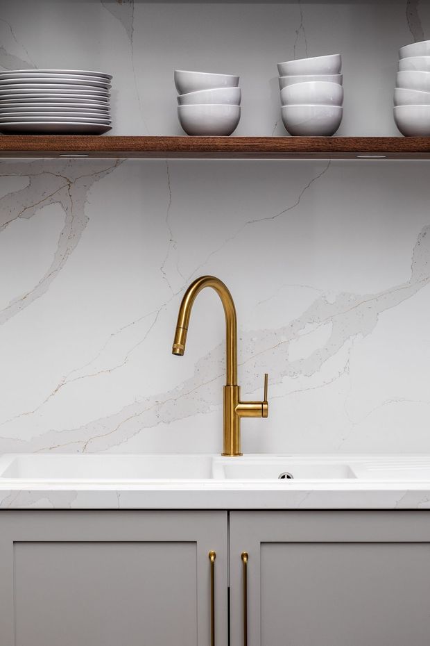 Close-up of a refined kitchen sink area with a white undermount sink, tall brushed-gold gooseneck faucet, grey shaker-style cabinetry with brass pulls, and a dramatic white marble-look veined splashback. Open timber shelving above displays neatly stacked white plates and bowls.
