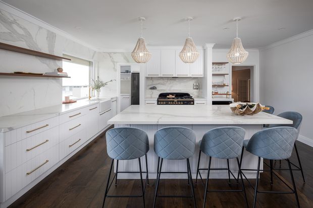 Bright, refined kitchen featuring extensive white cabinetry with slim brass-gold hardware, a large white marble-look island with four upholstered bar stools, dark timber flooring, marble-look wall surfaces, open display shelving, and three sculptural pendant lights. The room has a balanced, elegant contemporary-classic feel.