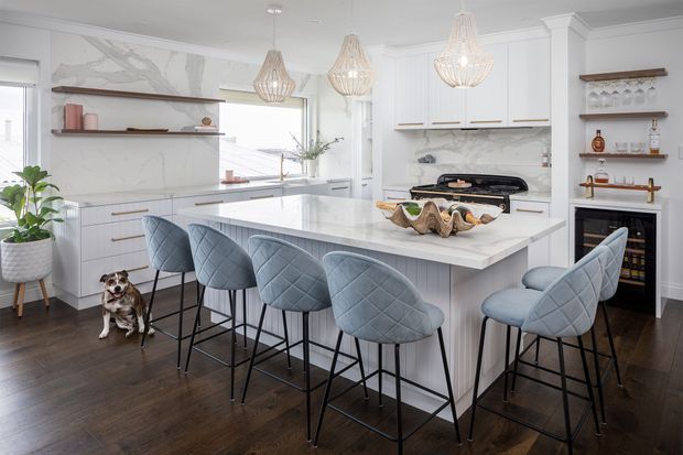Bright, spacious contemporary kitchen with a large white waterfall island, extensive white cabinetry, marble-look wall surfaces, open timber display shelving, and six upholstered blue-grey bar stools. Decorative pendant lights hang above the island, with dark timber flooring, integrated appliances, and a small wine refrigerator. A dog and potted plant are present but are not part of the architectural analysis.