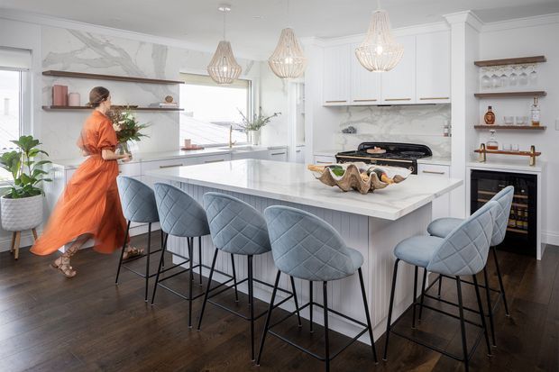 Bright, spacious contemporary kitchen with a large white waterfall island, integrated seating for several people, pale grey-blue upholstered bar stools, white cabinetry, marble-look splashback, dark timber flooring, open shelving, and three woven pendant lights. A small wine-storage area is built into the cabinetry at right. The room has a refined, airy coastal-inspired feel with strong contrast between the light fit-out and dark floor.