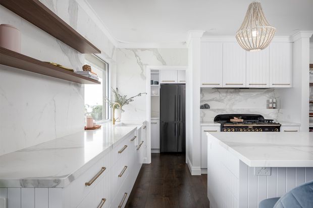 Bright, refined kitchen with white shaker-style cabinetry, veined marble-look wall surfaces and countertops, dark timber flooring, open timber shelving, a large island, and a statement woven pendant light. A black range cooker and integrated dark refrigerator provide contrast.
