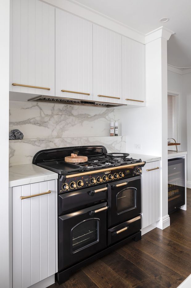Bright, refined kitchen focused on a large black range cooker with brass controls and handles. White vertically panelled cabinetry with brass bar pulls surrounds a dramatic veined marble backsplash and benchtop, while dark timber flooring adds contrast. A partial adjoining area is visible beyond the column.