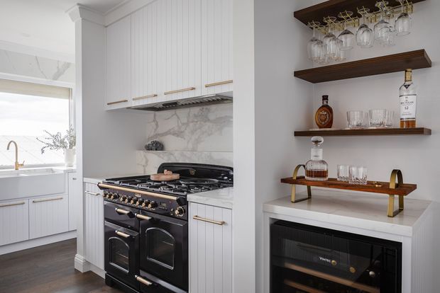 Elegant traditional kitchen with white vertical-panel cabinetry, brass hardware, a large black range cooker, and a marble-look splashback. To the right is a built-in drinks and wine area with dark timber floating shelves, glassware, spirits, and a wine fridge. The adjacent utility or sink area is visible through the opening on the left.