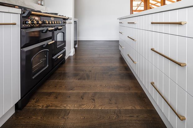 A low-angle view of a refined kitchen featuring a large black range cooker with brass controls and handles, white vertically panelled cabinetry, and a dark natural timber floor. A central island with long brass pulls dominates the right side, while white walls and timber-framed glazing are visible in the background.