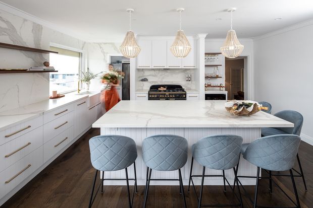 Bright, spacious kitchen with a large white marble-look island, four upholstered bar stools, extensive white cabinetry, integrated range cooking area, open shelving, and three woven pendant lights. Dark timber flooring and pale blue-grey seating add contrast to the refined, contemporary fit-out.
