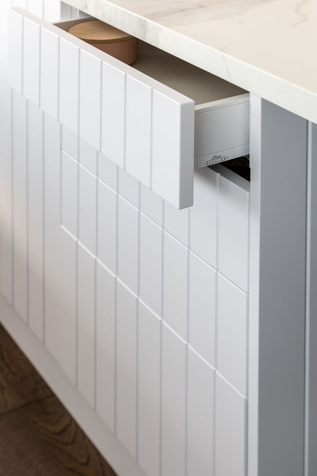 Close-up detail of white built-in kitchen cabinetry with vertically fluted or panelled drawer fronts. One drawer is open, revealing an internal storage compartment and a round timber container, while a pale stone benchtop sits above. Dark timber flooring is visible below.
