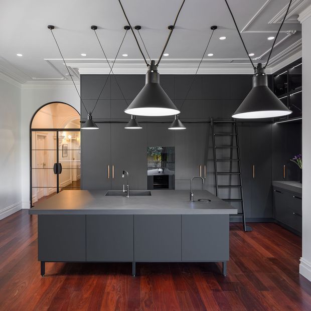 A sophisticated contemporary kitchen with a large dark island, integrated sink and tapware, extensive floor-to-ceiling charcoal cabinetry, and a grey benchtop. Multiple black pendant lights hang from a coffered and moulded white ceiling. The room features rich reddish-brown timber flooring, black-framed glazing and an arched opening leading to an adjoining space, with a ladder integrated against the cabinetry.