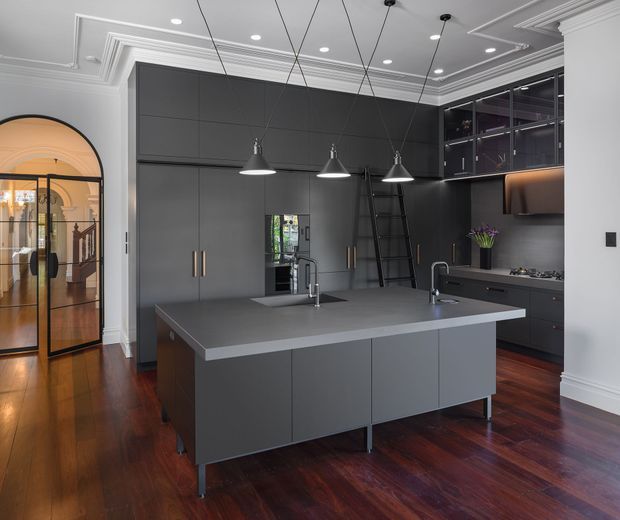 A sophisticated dark-toned kitchen with a large central island, integrated sink and cooktop, extensive floor-to-ceiling cabinetry, open display shelving, and three suspended pendant lights. The room features rich dark timber flooring, charcoal cabinetry and benchtops, white decorative ceiling mouldings, and a glazed arched doorway leading to an adjoining space.