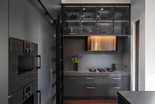 A compact, dark-toned contemporary kitchen with charcoal cabinetry, integrated appliances, a grey stone-look benchtop and splashback, black-framed glass-front overhead cupboards, and warm metallic accents. A sculptural illuminated rangehood feature sits above the cooktop, with a narrow window and a dark island or peninsula visible at the right.