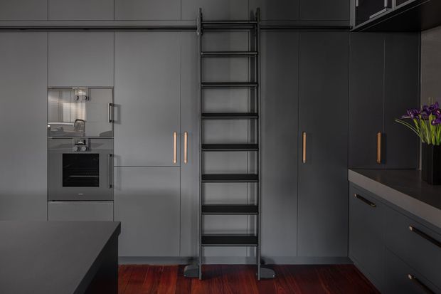 A sophisticated dark-toned contemporary kitchen with full-height matte cabinetry, integrated stainless-steel oven, central rolling library ladder, dark benchtops, and deep reddish timber flooring. Minimal brass-toned handles and a vase of purple flowers provide subtle accents.