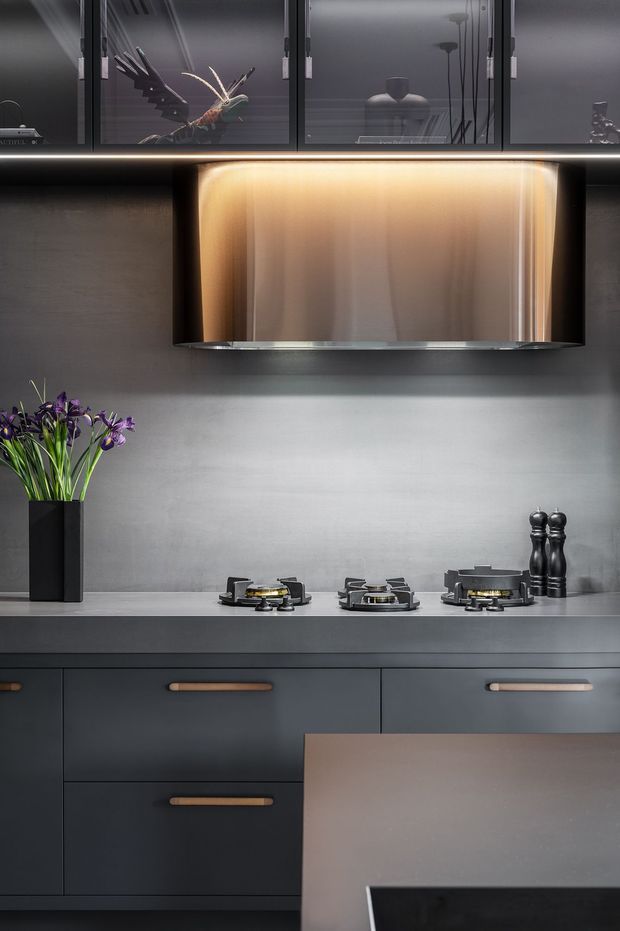 A refined dark-toned kitchen vignette featuring a black gas cooktop beneath a large illuminated cylindrical rangehood. Matte charcoal cabinetry with warm timber-look handles lines the lower half, while a smooth grey splashback and matching benchtop create a monochromatic backdrop. Black-framed glass display cabinets above contain decorative objects, and a small vase of purple flowers adds contrast.