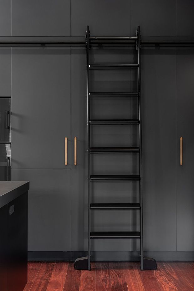 A dark, contemporary kitchen with floor-to-ceiling matte charcoal cabinetry, integrated handles, and a prominent black rolling library-style ladder positioned against the upper cabinets. A dark island edge is visible at left, while reddish-brown timber flooring adds warmth and contrast.
