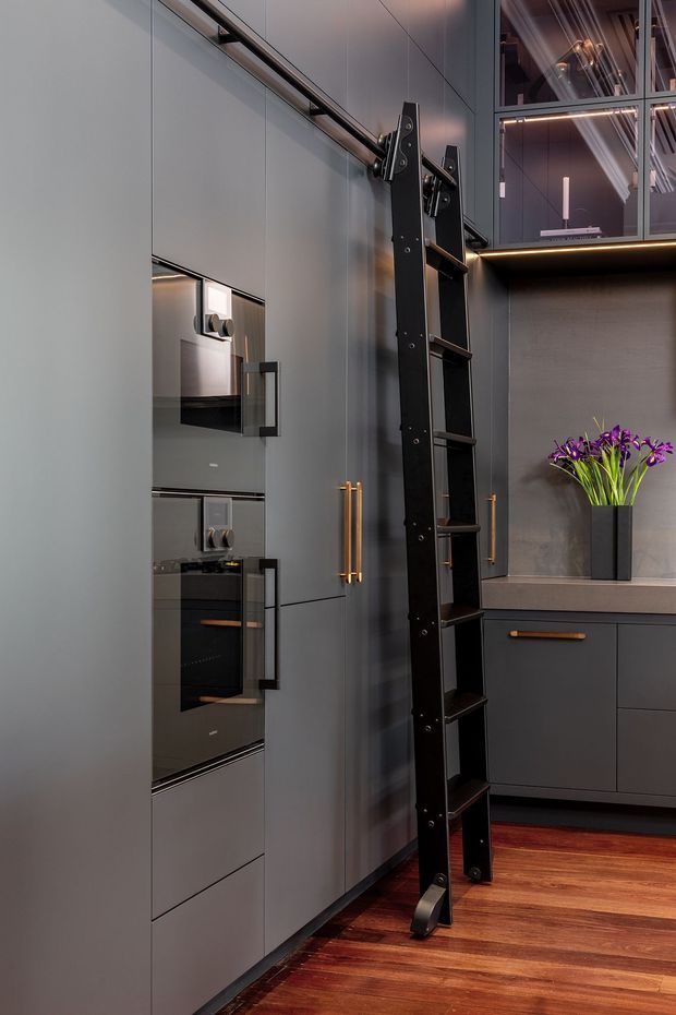 A sophisticated, dark-toned kitchen or compact living space with floor-to-ceiling matte grey cabinetry, integrated appliances, slim bronze handles, and a tall black sliding ladder providing access to upper storage. A dark stone or plaster feature wall and built-in cabinetry frame a vase of purple flowers, while warm timber flooring and concealed under-cabinet lighting add contrast. Glazed upper cabinetry reflects the room and contributes to the refined, contemporary atmosphere.