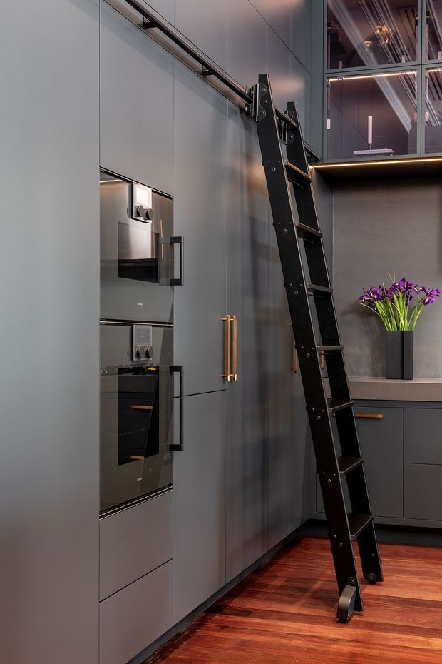 A sophisticated contemporary kitchen with full-height matte dark-grey cabinetry, integrated appliances, brass-gold handles, and a distinctive black rolling ladder reaching upper storage. Warm timber flooring contrasts with the cool cabinetry, while a dark stone-look benchtop and backsplash frame a built-in display area with under-cabinet lighting and purple flowers. A glass-fronted upper cabinet adds visual depth.