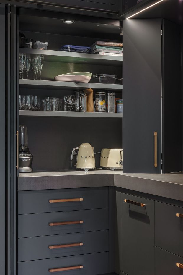 A compact, dark-toned kitchen pantry or appliance-storage zone with matte charcoal cabinetry, open shelving, integrated LED lighting, and warm timber or copper-toned handles. Shelves contain glassware, dishes, jars, and small appliances, while grey stone-look benchtops wrap around the corner.