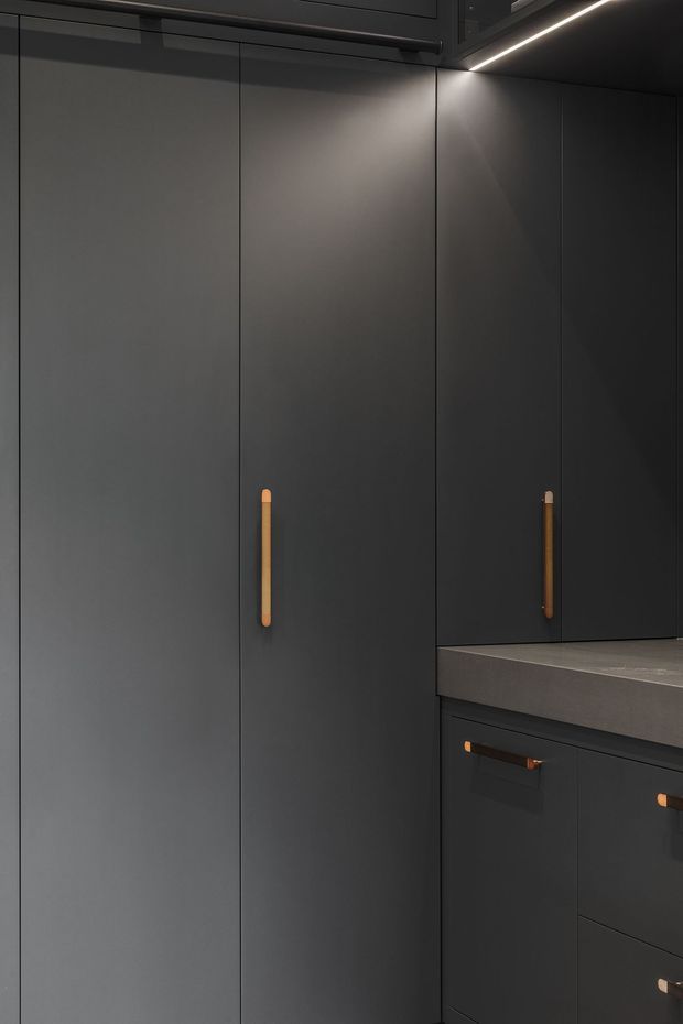 Close-up of a dark, minimalist kitchen fit-out featuring floor-to-ceiling matte charcoal cabinetry, integrated upper cabinets, slim warm timber-toned handles, and a dark grey stone-look benchtop. Concealed linear LED lighting highlights the cabinetry and work surface.