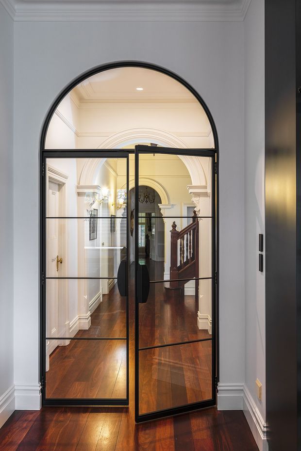 Interior view of a refined hallway or entry passage with dark polished timber flooring, white decorative wall and ceiling mouldings, and a black-framed arched glazed door. The door opens toward a classical corridor with arched architectural details, a chandelier, and a timber staircase with turned balusters.