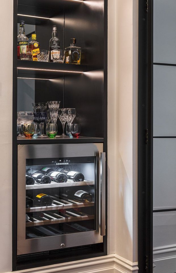 A compact built-in home bar cabinet with black illuminated shelving displaying liquor bottles and crystal stemware, above an integrated stainless-steel wine refrigerator. The unit is set into a light painted wall with black-framed glazed cabinetry or door visible at right, creating a refined, moody bar storage area.