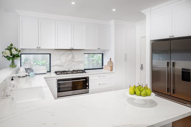 Bright, spacious white kitchen with extensive shaker-style cabinetry, a large marble-look island benchtop, integrated stainless-steel appliances, gas cooktop, and veined splashback. Recessed ceiling lights and decorative cornices give the room a refined, classic appearance, while windows bring in greenery and natural light.