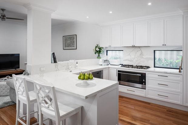 Bright, refined white kitchen with a large marble-look island, integrated seating, extensive shaker-style cabinetry, stainless steel appliances, gas cooktop, and timber flooring. The open-plan living area is partially visible beyond the kitchen, with aquarium-like glazed features and decorative styling.