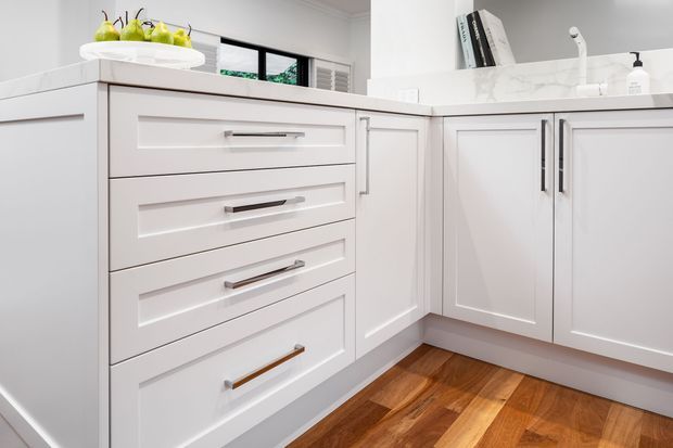 Bright, compact white kitchen with Shaker-style cabinetry, recessed-panel drawers and doors, slim dark hardware, a pale benchtop, and warm timber flooring. A small window and open shelving add light and functionality.