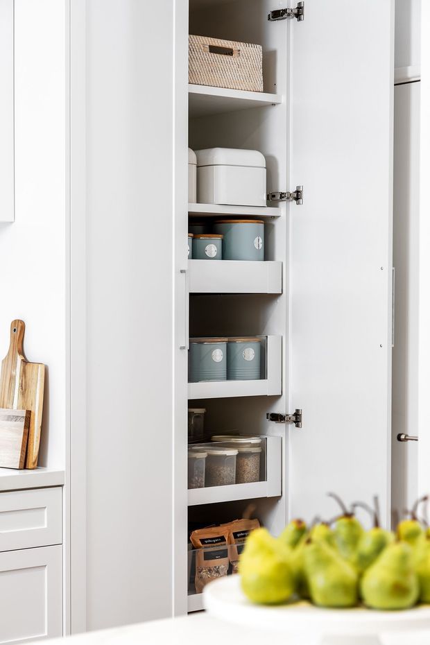 Close-up view of a tall white kitchen pantry cabinet with the door open, showing multiple adjustable shelves and pull-out drawers containing storage boxes, canisters, and pantry goods. Part of the surrounding white kitchen cabinetry and timber chopping boards are visible, with a blurred bowl of fruit in the foreground.