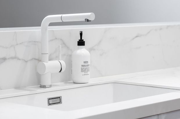 Close-up of a minimalist white kitchen sink with a sculptural white mixer tap, integrated basin, white cabinetry, and a white marble-look splashback with subtle grey veining. A soap dispenser sits beside the tap. The composition is clean, bright, and monochromatic.