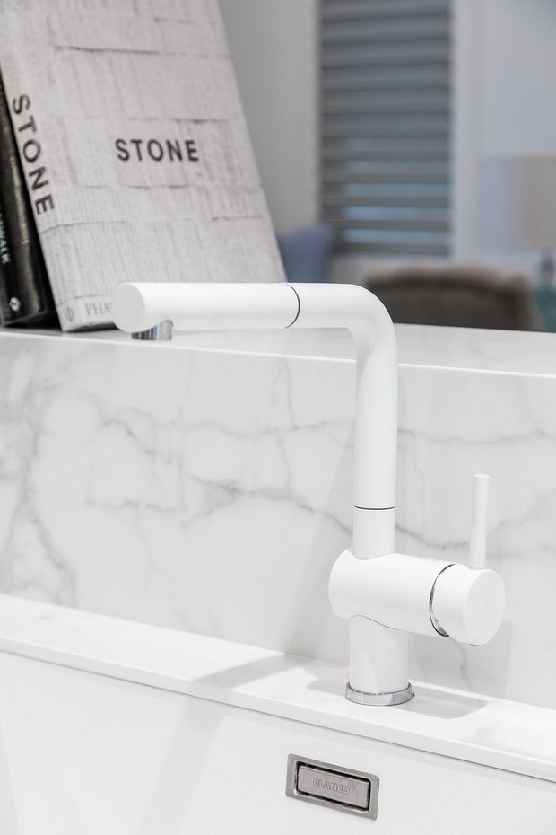 Close-up product-style photograph of a contemporary white kitchen mixer tap mounted over a white sink. The sink area features a white marble-look splashback with subtle grey veining and a matching white benchtop. A design book and blurred interior background are visible behind the tap.