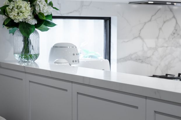 Bright contemporary kitchen featuring a long white island with integrated grey cabinetry and slim recessed handles, a white marble-look splashback, dark-framed internal glazing, and a minimalist white appliance. A floral arrangement in a ribbed glass vase adds a soft decorative accent.
