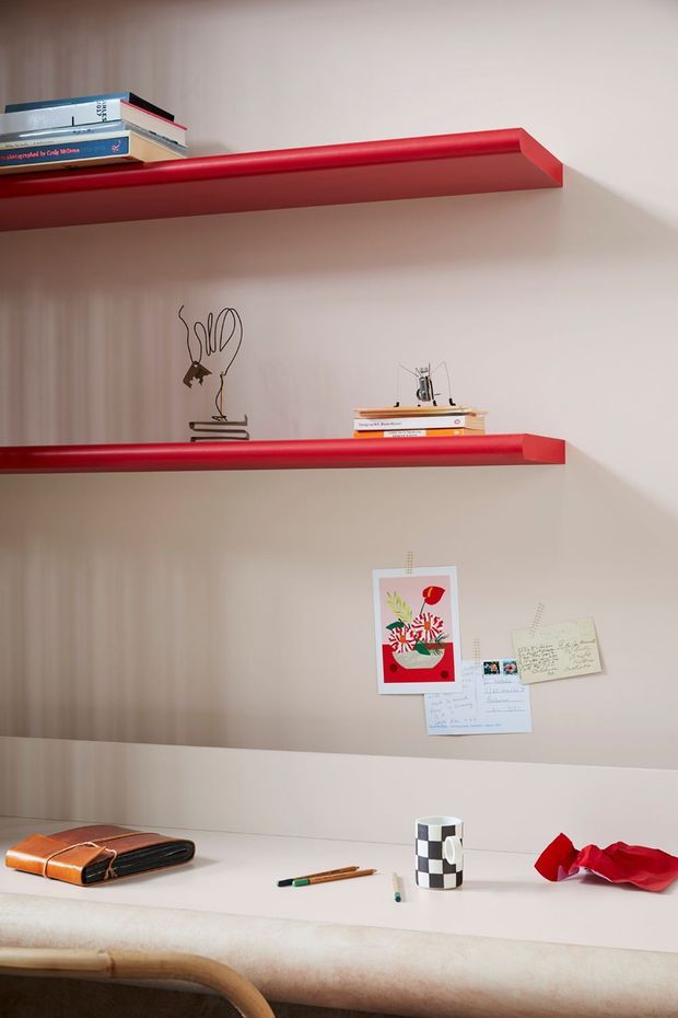 A close-up of a stylish built-in study nook or home office with pale wood wall panelling, two bold red floating shelves, books, small decorative objects, pinned notes and artwork, and a light-coloured desk surface. A black-and-white checkered mug, stationery, wallet and red fabric accent sit on the desk, with part of a curved timber chair visible in the foreground. The composition has a playful, curated contemporary feel.