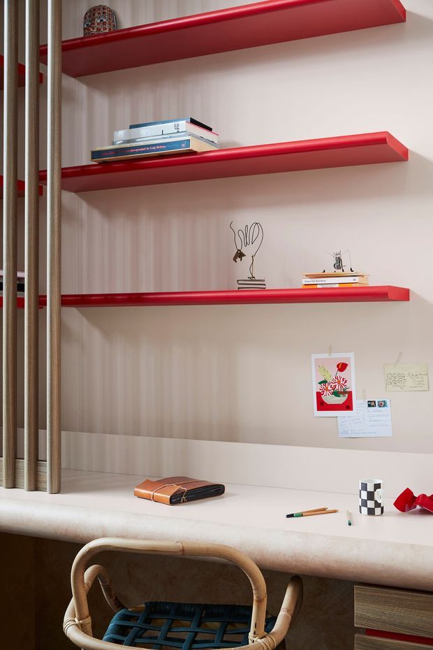 A compact built-in study nook with a pale wood-panelled backdrop, three bold red floating shelves displaying books and small objects, and a light stone or composite desk surface. A woven timber-framed chair sits at the desk, which includes dark wood cabinetry and drawers below. The space has a playful, contemporary, highly curated character with warm natural textures and strong red accents.