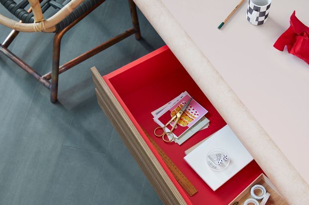 Top-down detail of an open bright-red drawer in a light timber built-in cabinet or desk, containing stationery and craft items. A woven timber-framed chair sits on a smooth dark grey floor beside the joinery, while a pale countertop or work surface with pencils, a patterned cup, and red paper occupies the upper right.